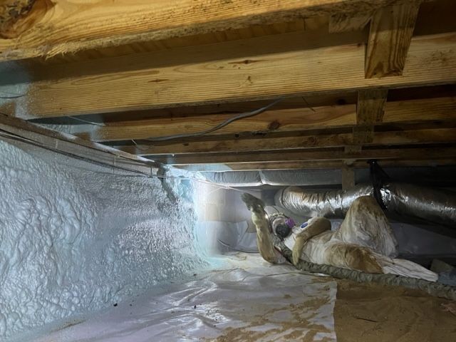 Person in protective gear working in insulated crawl space with wooden beams and ventilation ducts overhead.