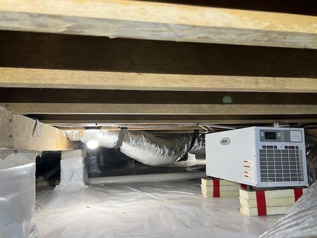 Crawl space with vapor barrier and dehumidifier unit, exposed wooden beams above.